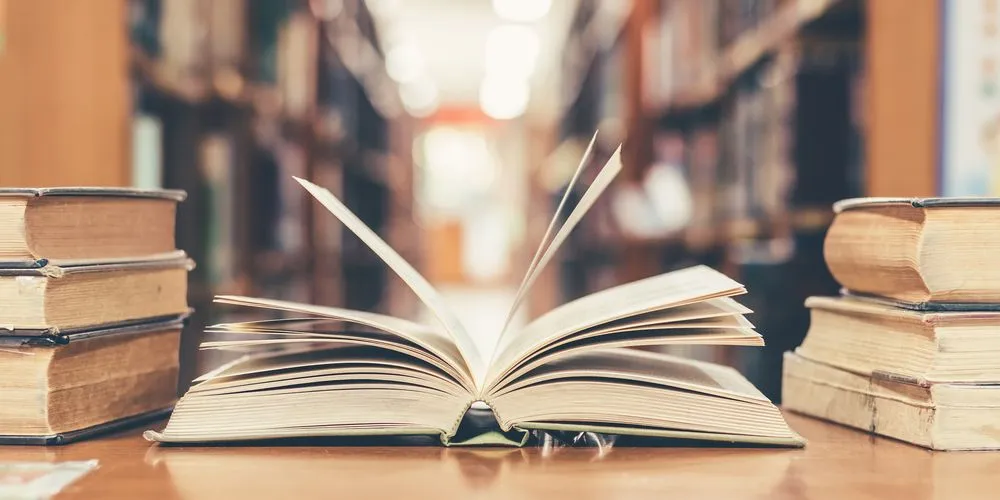 Open book with flipping pages on wooden table, surrounded by stacked old books, blurred library shelves in background.