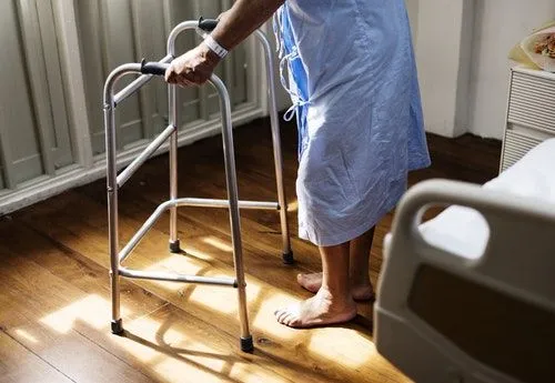 Elderly person in a blue hospital gown using a walker on wooden floor.