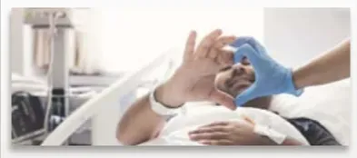 Patient lying in hospital bed making a heart shape with gloved healthcare worker's hands.