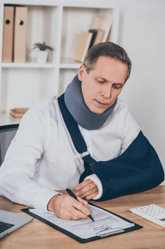 Man wearing a neck brace and arm sling, filling out a form at a desk with office supplies.