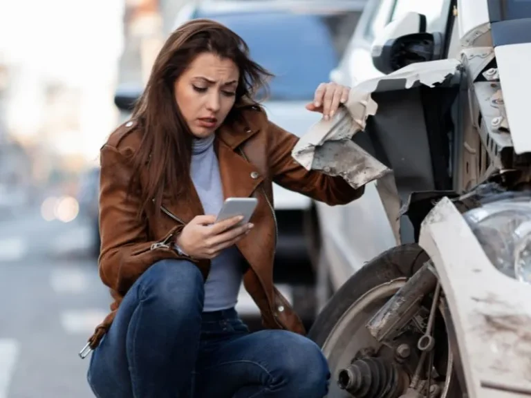 Woman in brown jacket examining damaged white vehicle while using smartphone.