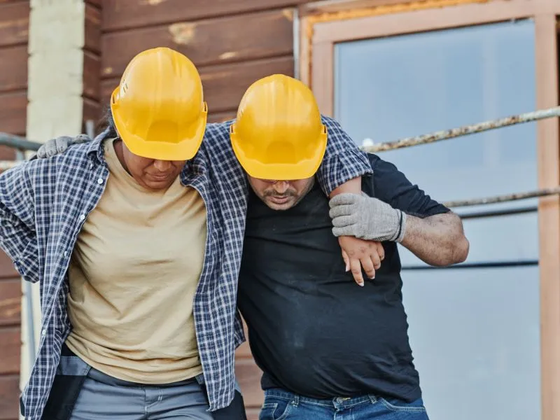 Two construction workers wearing yellow hard hats, one assisting the other.