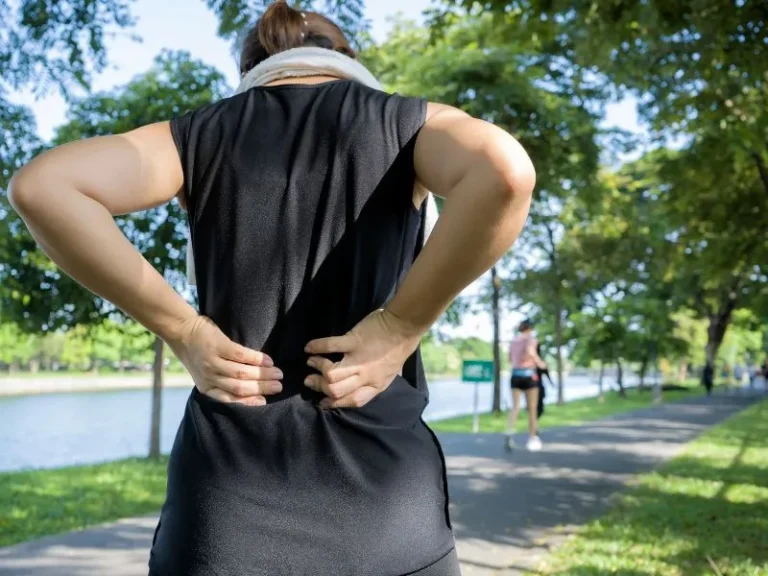 Person in black athletic wear holding lower back, standing on a shaded park pathway beside a river.
