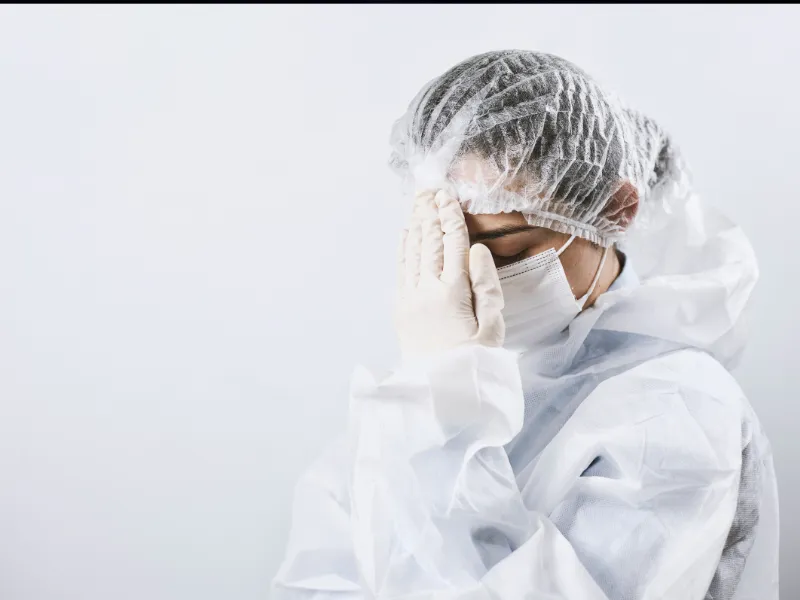 Healthcare worker in full protective gear, including mask and gloves, appearing stressed.