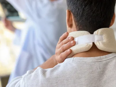 Man wearing white neck brace, holding neck with hand, person in background.