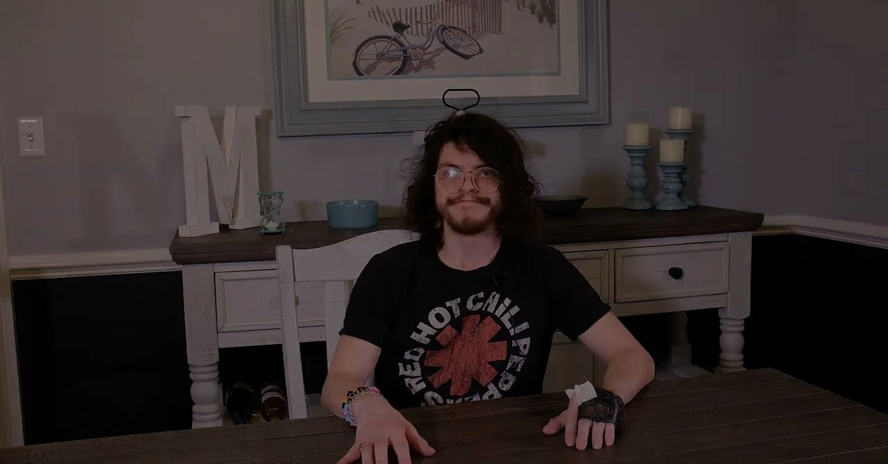 Young man with glasses and dark hair sitting at a wooden table in a dimly lit room.