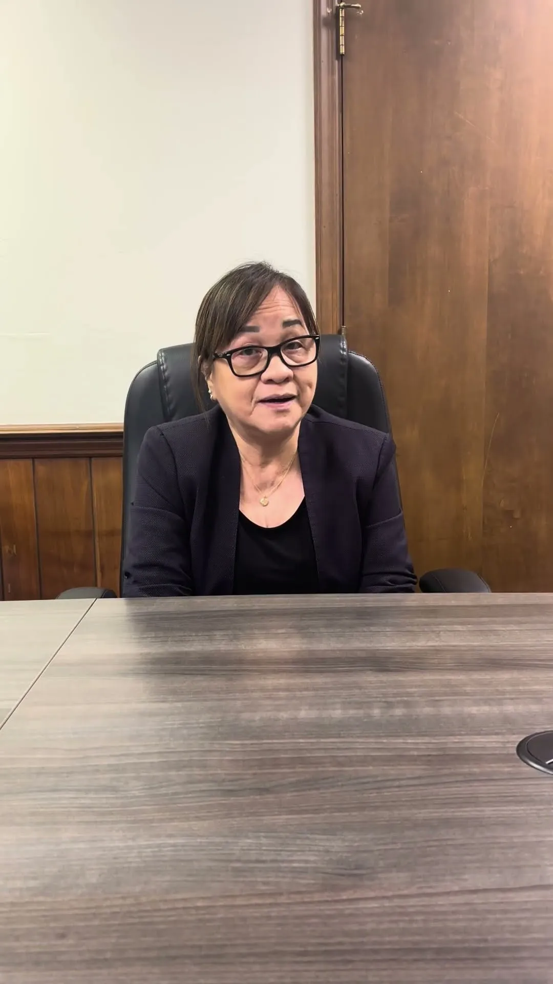 Middle-aged woman with glasses and dark blazer seated at a wooden conference table.