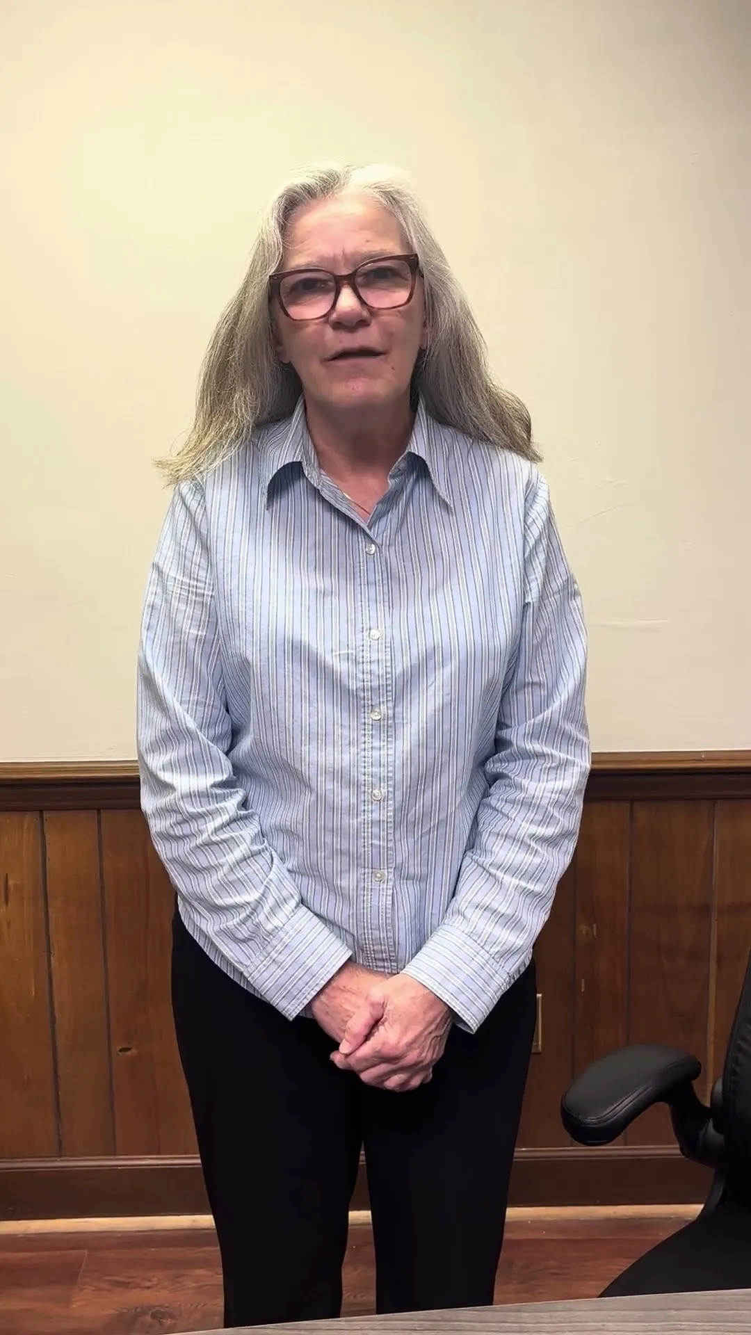 Middle-aged woman with long gray hair and glasses standing in office, wearing striped shirt and black pants.