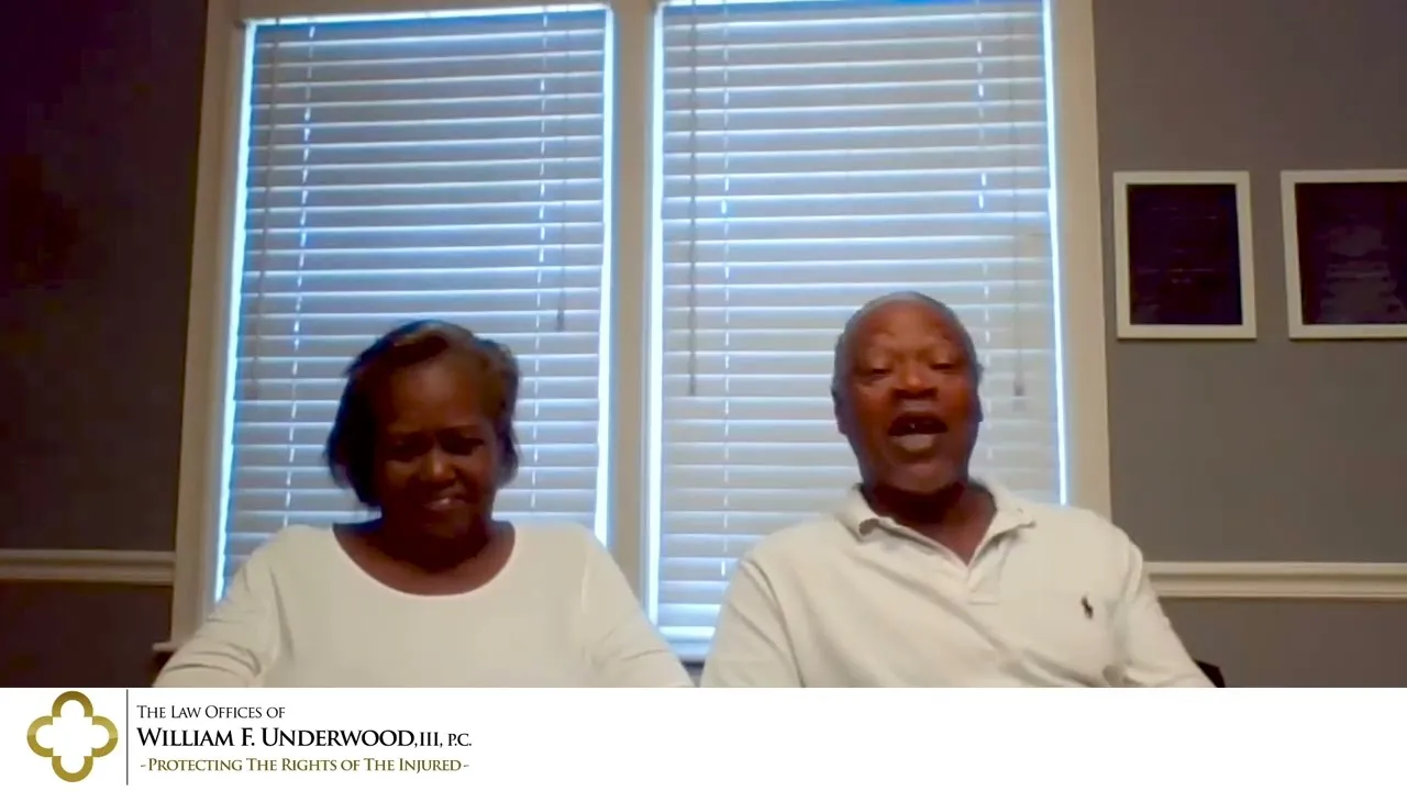 Two middle-aged individuals seated indoors in front of a window with closed blinds, wearing white shirts.