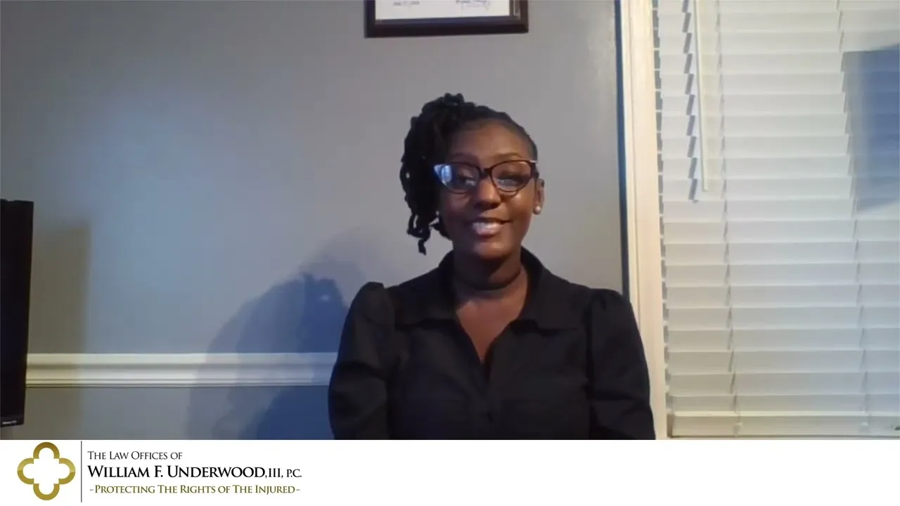 Professional woman with glasses and braided hair, seated in office with gray walls and white blinds.