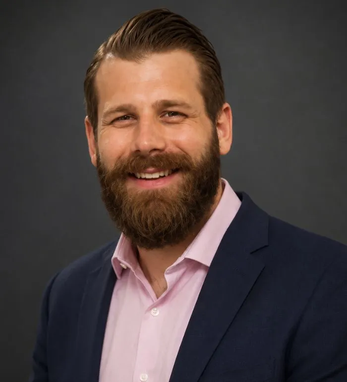 Portrait of a smiling man with a beard, wearing a navy blazer and light pink shirt.
