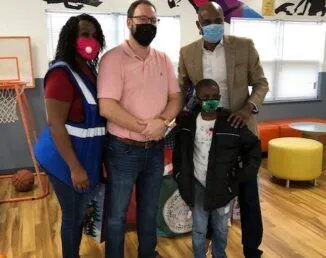 Four people wearing face masks stand indoors on a wooden floor with colorful furniture and a basketball hoop in the background.