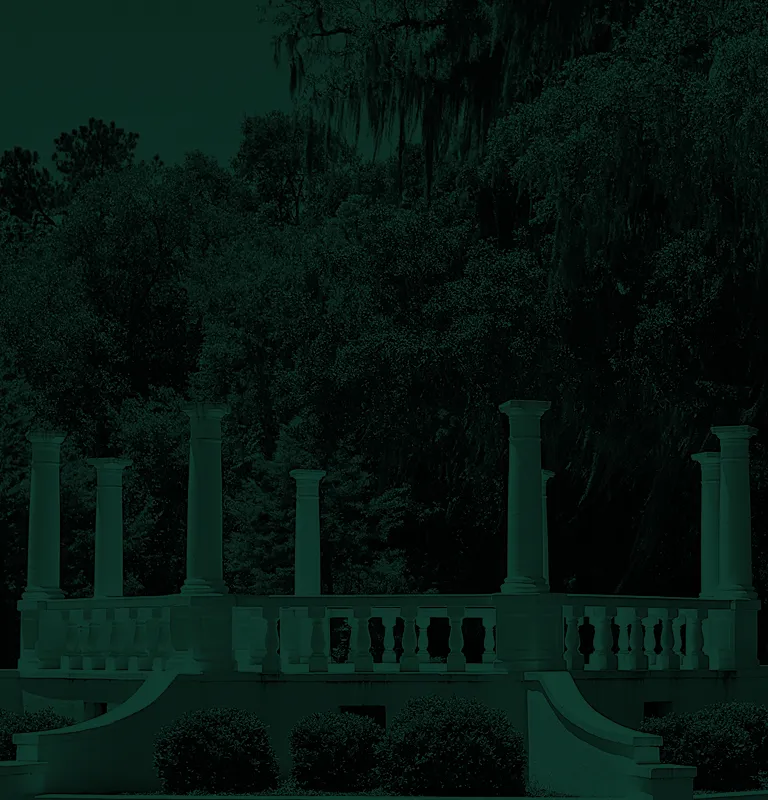 White classical gazebo with columns and balustrades surrounded by green trees and bushes.