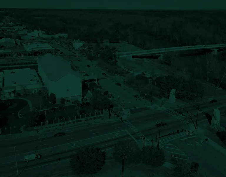 Aerial view of a suburban intersection with vehicles, buildings, and a nearby bridge over a wooded area.