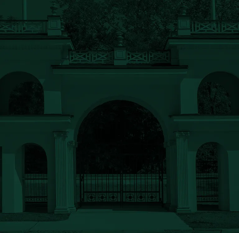 White classical archway with columns and decorative metal gate, surrounded by trees.
