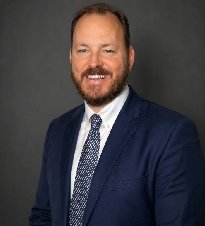 Professional portrait of a smiling man with beard wearing navy suit and patterned tie against gray background.