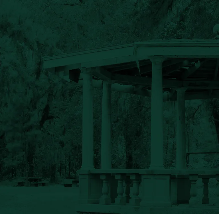 Stone gazebo with columns in a wooded park area, surrounded by trees and benches.