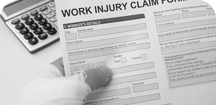 A hand with a bandaged thumb filling out a work injury claim form next to a calculator.