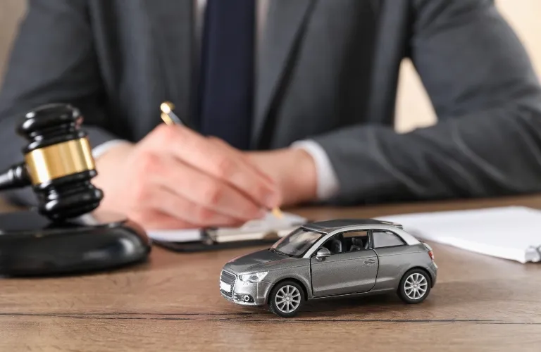 Gray toy car on wooden table with judge's gavel and person writing in background.