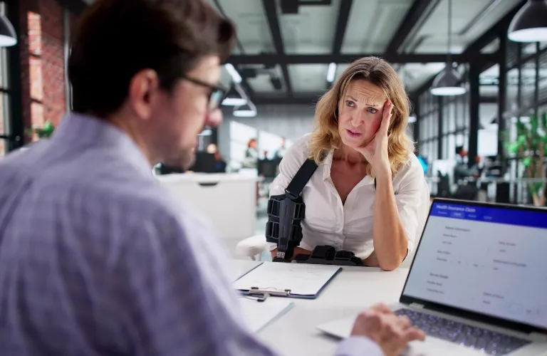 Two professionals in an office; woman with arm brace appears concerned, man using laptop.