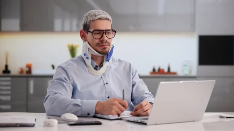 Man with neck brace and glasses working on laptop in modern kitchen setting.