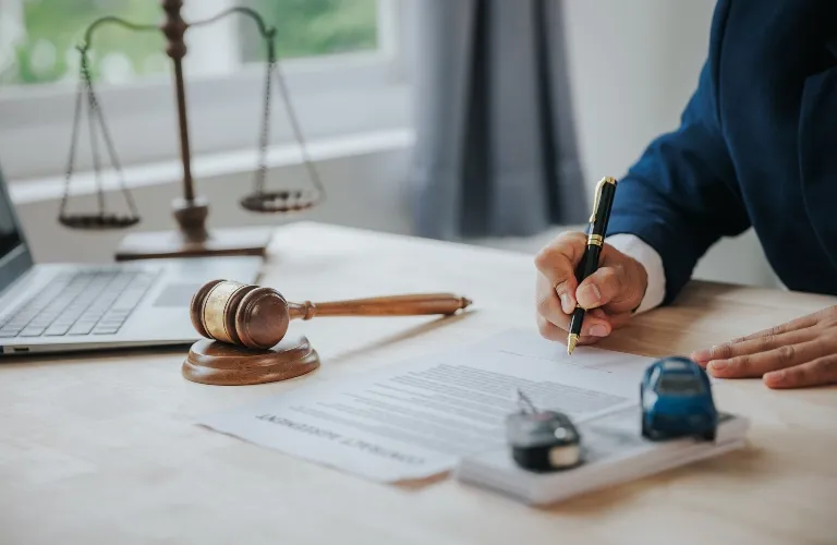 Person in blue suit signing legal document on desk with gavel, scales, and laptop.