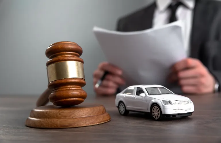Wooden judge's gavel, white toy car, and person in suit holding papers on desk.