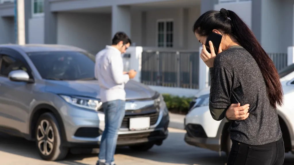 Woman in gray top on phone and man in white shirt inspecting two cars after collision.