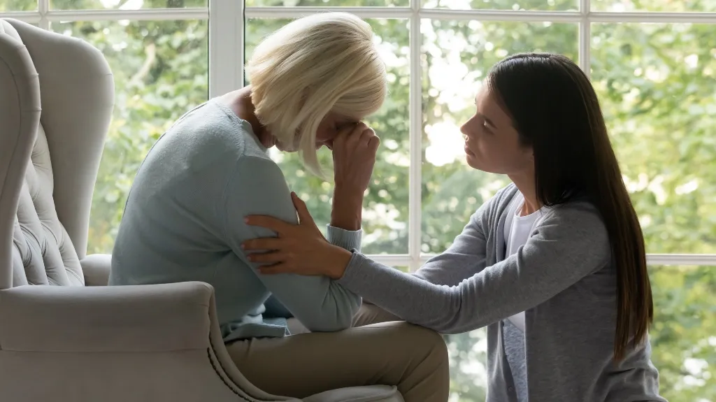 Two women in a bright room by a window, one comforting the other who appears distressed.
