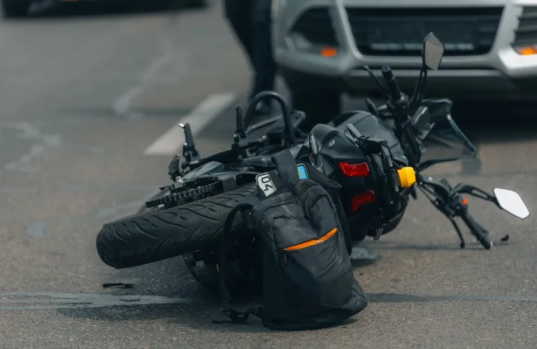 Black motorcycle lying on its side on asphalt road with a black backpack beside it.