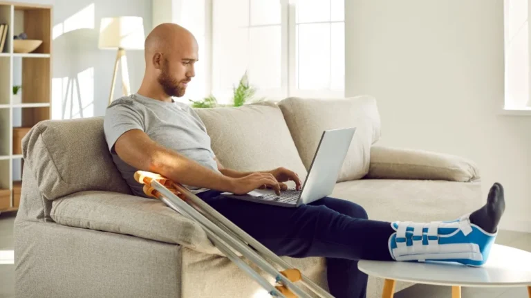 Man with leg cast and crutches working on laptop while seated on beige sofa.