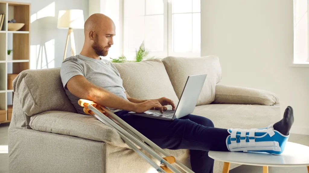 Man with leg cast and crutches working on laptop while seated on beige sofa.