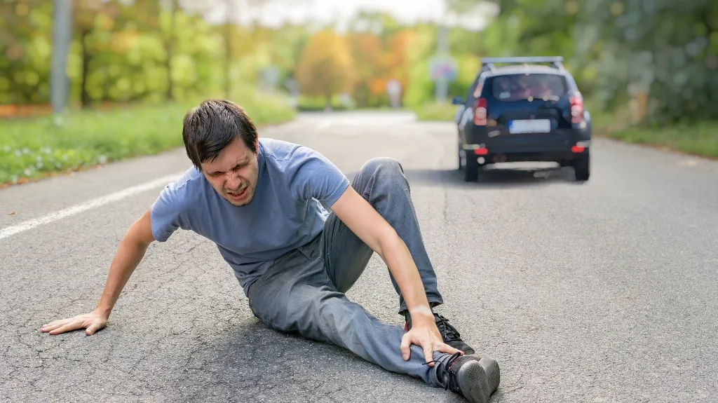 Man in gray shirt and pants sitting on road, holding injured leg, with black car in background.