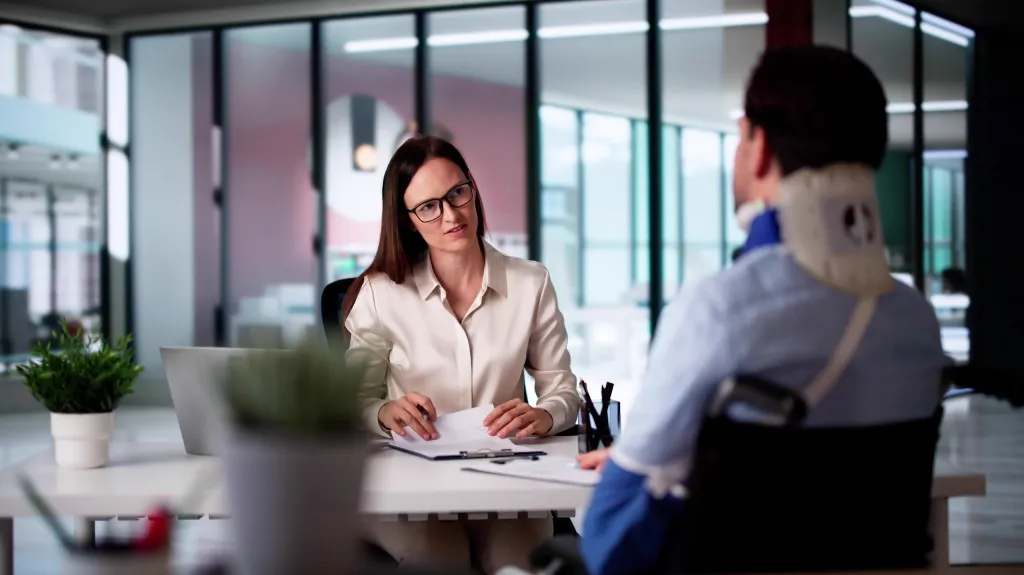 Professional woman with glasses conducting an office consultation with an injured man wearing a neck brace.