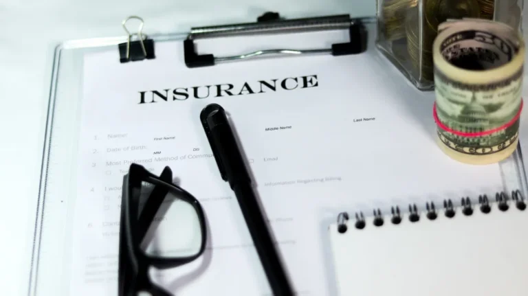 Insurance form on clipboard with black pen, glasses, rolled cash, and notebook.