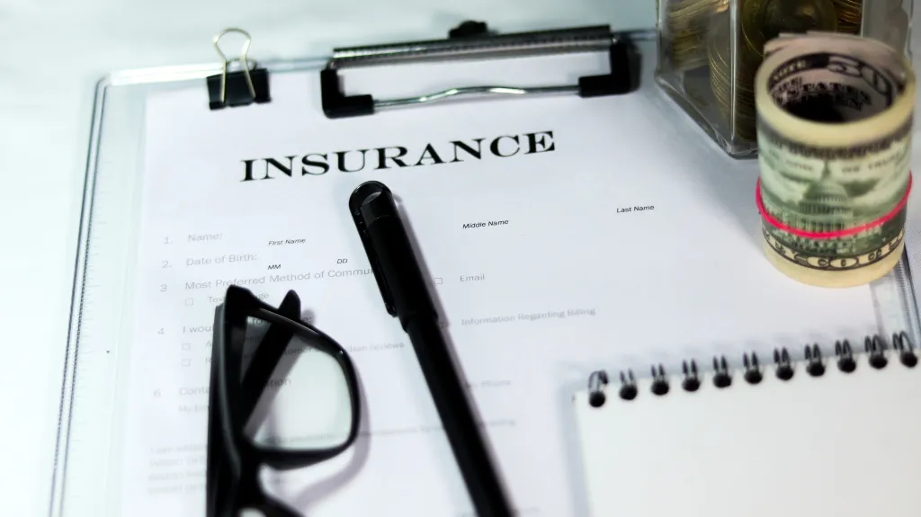 Insurance form on clipboard with black pen, glasses, rolled cash, and notebook.