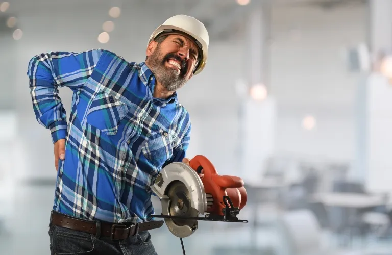 Man in blue plaid shirt and white hard hat holding circular saw, grimacing in pain, holding his lower back.