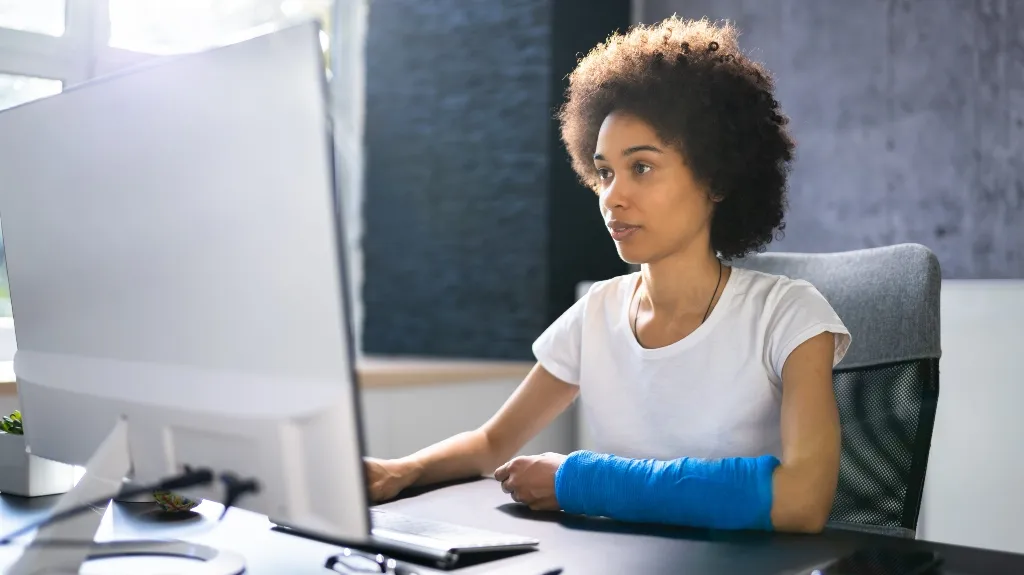 Young woman with a blue arm cast working at a computer in a modern office.