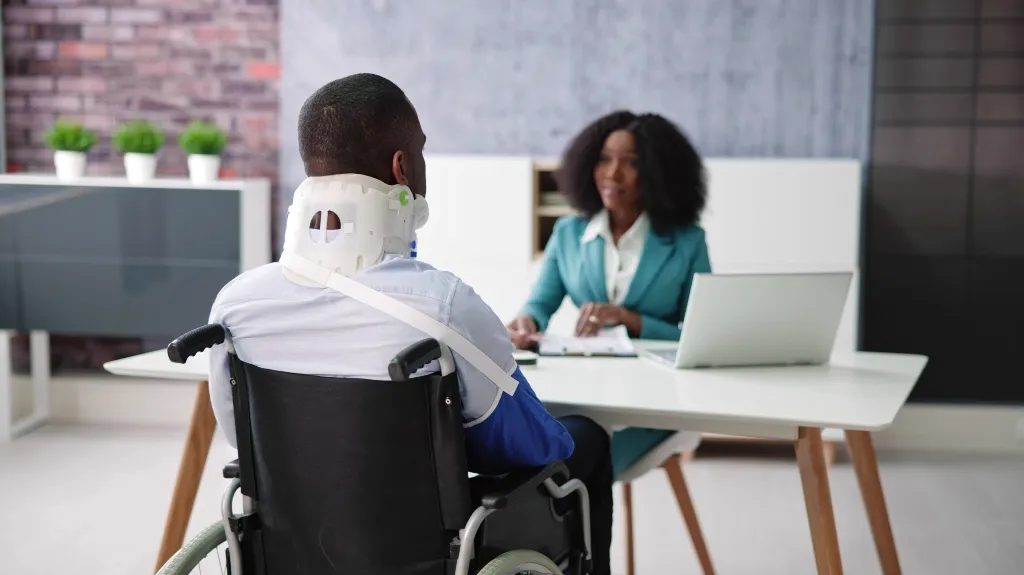 Man in wheelchair with neck brace consulting professional woman in teal suit at office desk.