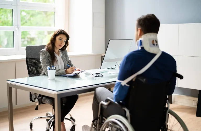 Professional woman consulting with injured man in wheelchair wearing a neck brace in an office.