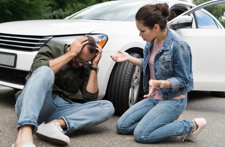 Man sitting on ground holding head, woman kneeling beside him near white car.