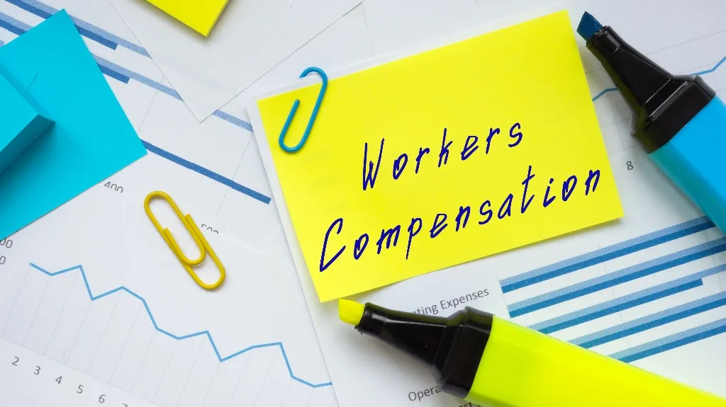 Yellow sticky note with "Workers Compensation" text, surrounded by charts, paperclips, and highlighters.