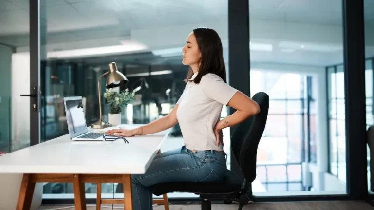 Young woman in white shirt and jeans sitting at desk, holding her lower back in discomfort.