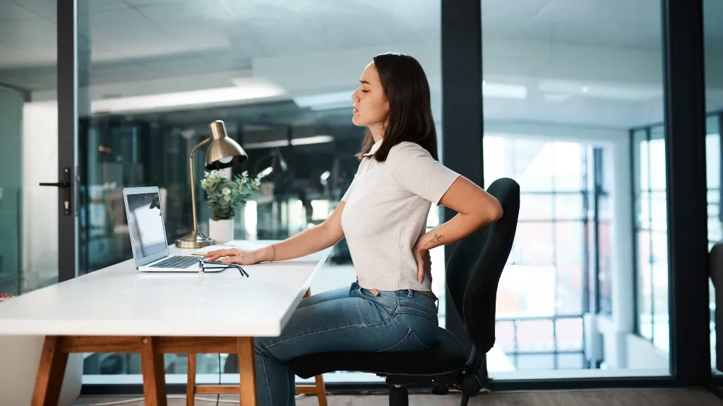 Young woman in white shirt and jeans sitting at desk, holding her lower back in discomfort.