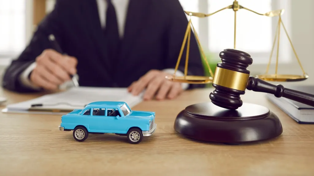 Close-up of a blue toy car, wooden judge's gavel, and golden scales of justice on desk.