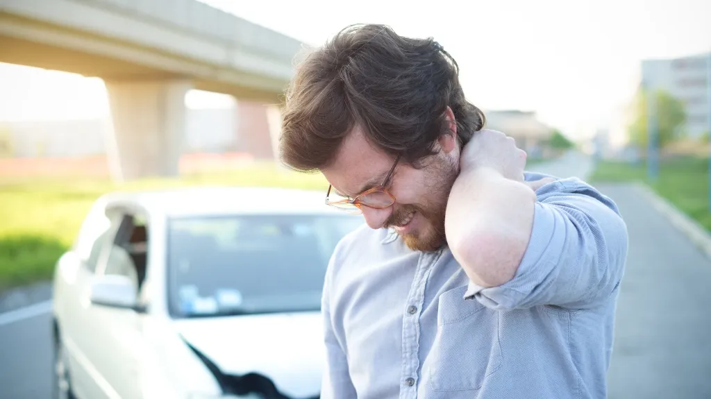 Man in light blue shirt holding neck in pain, standing beside damaged white car.