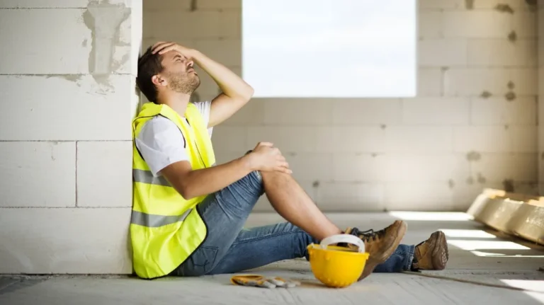Construction worker in yellow safety vest sitting on floor, holding head, next to yellow hard hat.
