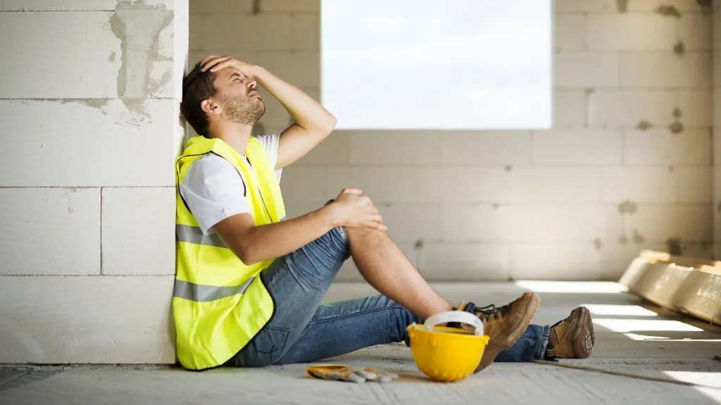 Construction worker in yellow safety vest sitting on floor, holding head, next to yellow hard hat.